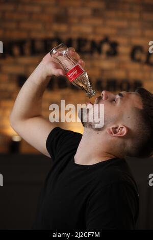 RIVNE, UKRAINE JUNI 26 2020: Junger bärtiger Mann trinkt Coca Cola aus der Flasche im Friseurladen auf Backsteinmauer Hintergrund. Stockfoto