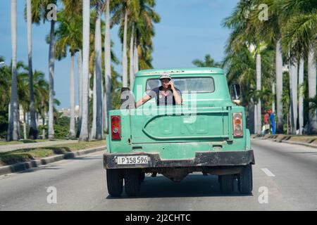 Der junge Kubaner fährt auf der Rückseite eines Pick-up-Trucks durch eine Straße in Havanna, Kuba telefoniert. Stockfoto