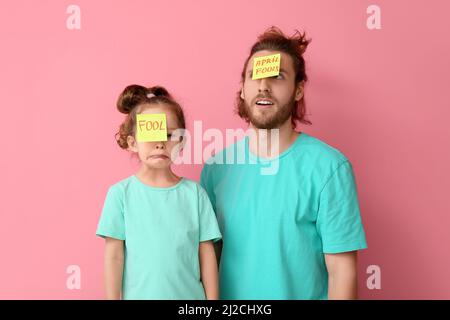 Lustiger Mann und ihre kleine Tochter mit Haftnotizen auf ihren Stirnen vor rosa Hintergrund. Das Fest des April-Narren Stockfoto