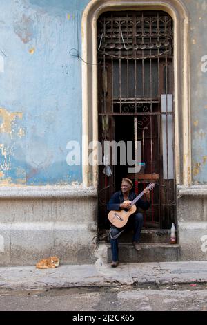 Älterer Mann singt und spielt auf einer Straße Gitarre, während seine Katze in Havanna, Kuba, auf dem Boden lauscht. Stockfoto