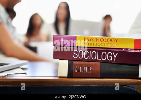 Sozialstudien. Ausgeschnittene Aufnahme von Universitätslehrbüchern auf einem Schreibtisch mit verschwommenen Studenten im Hintergrund. Stockfoto