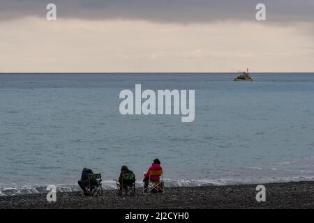 Drei Menschen von hinten sitzen am Strand am Meer mit einem Fischerboot an einem stürmischen Tag, Bordighera, Imperia, Ligurien, Italien Stockfoto