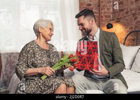 Ein Unternehmer mittleren Alters, der den Geburtstag seiner Mutter feiert und ihr einen Strauß roter Tulpen und ein Geschenk gibt. Hochwertige Fotos Stockfoto