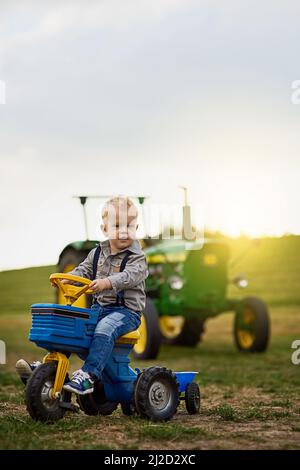 Es gibt wenig, was man nicht über das Leben auf dem Bauernhof lieben kann. Porträt eines entzückenden kleinen Jungen, der auf einem Spielzeugauto auf einer Farm reitet. Stockfoto