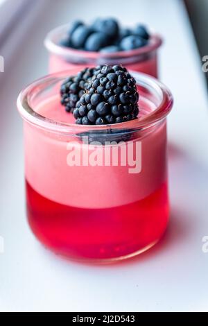Eine Nahaufnahme eines Brombeerjello-Joghurts-Dessert in durchsichtiger Glasschale auf weißem Tisch mit Heidelbeer-Jello im unscharfen Hintergrund Stockfoto
