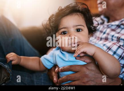 Versuchen herauszufinden, was los ist. Porträt eines fröhlichen kleinen Jungen, der auf dem Schoß seines Vatis sitzt, während er während des in die Kamera schaut Stockfoto