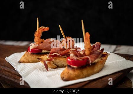 Bruschettas mit serrano-Schinken, Kirschtomaten und gegrillten Garnelen auf einem Holzbrett. Mediterranes Speisekonzept. Stockfoto
