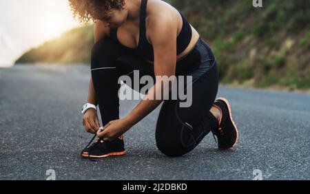 Schnüren Sie sich, damit Sie nicht über Ihre Ziele stolper. Aufnahme einer sportlichen jungen Frau, die ihre Schnürsenkel beim Training im Freien bindet. Stockfoto