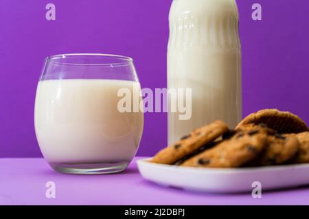 Nahaufnahme einer Glasflasche und eines großen Glases mit Milch und einigen süßen Schokoladenstückchen in einer violetten oder violetten Umgebung. Speicherplatz kopieren. Stockfoto