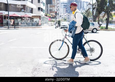Latino-Mann mit Brille in legerer Kleidung, der mit seinem Fahrrad mit roter Mütze, gelben Kopfhörern und einem Rucksack die Straße entlang in der Stadt läuft Stockfoto
