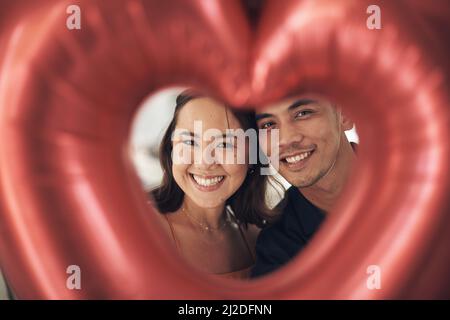 Stellen Sie sich das perfekte Paar vor. Aufnahme eines jungen Paares, das zu Hause durch einen herzförmigen Ballon posiert. Stockfoto