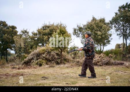 Männlicher Jäger in Camouflage-Oberbekleidung und Mütze, der eine Waffe trägt, die auf dem Feld läuft Stockfoto