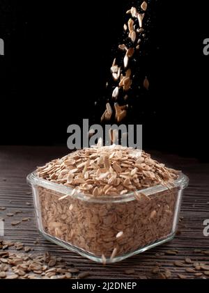 Haferflocken oder rohes Haferflocken fallen in einen quadratischen Glasbehälter auf schwarzem Hintergrund und auf einen Holztisch. Glasbehälter voll rohem Haferflocken. Stockfoto