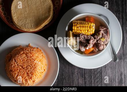 Mexikanisches Essen, Rinderbrustbrühe mit Gemüse auf einem weißen Teller mit Zitronenscheiben und mexikanischem Reis mit Tortillas auf einem alten Holztisch. Stockfoto