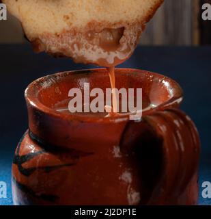 Süßes Brot in eine mexikanische Tasse heiße Schokolade oder Kakao auf einem blauen Tisch und einem Holzhintergrund eintauchen. Makrofotografie. Stockfoto