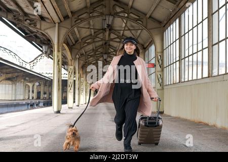 Junge süße Mädchen mit Hund und Koffer auf Plattform wartet auf Zug in rosa Mantel, am Bahnhof Vitebsk in St. Petersburg, Russland. Reisender Teenager mit Hund Stockfoto