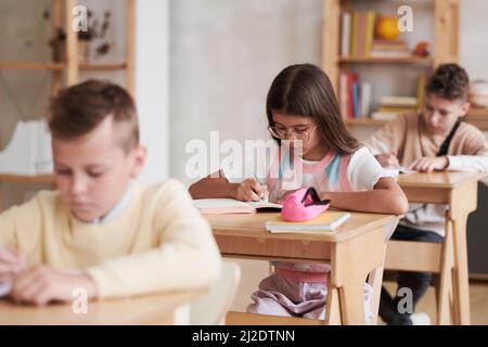 Eine Gruppe von Kindern, die im Klassenzimmer an Tischen in Reihe sitzen, konzentriert sich auf Schülerinnen, die eine Brille tragen Stockfoto