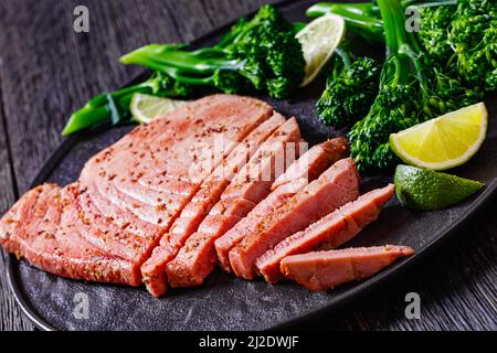 Nahaufnahme eines gebratenen Thunfischsteaks mit gekochtem Brokkolini und Limette auf schwarzem Teller auf dunklem Holztisch, horizontale Ansicht von oben Stockfoto