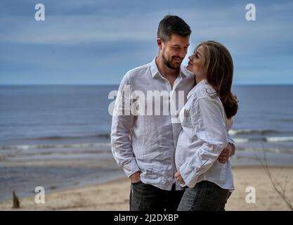 Ein Paar in passender Kleidung steht am Meer, umarmt sich mit Liebe und schaut mit einem glücklichen Lächeln. Die Frau ist schwanger. Stockfoto