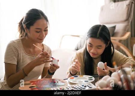 Happy Family Time on Oster Holiday Konzept, attraktive asiatische Frau mit ihrer Schwester in einem Wohnzimmer, malen ein Osterei zusammen. Stockfoto