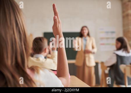 Nahaufnahme eines unerkennbaren Schulmädchen, das im Unterricht die Hand hebt, mit einer Lehrerin im Hintergrund, Platz kopieren Stockfoto