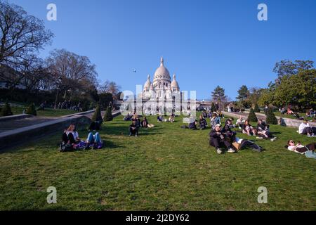 Paris, Frankreich - 22. März 2022 : die Basilika des Heiligen Herzens von Paris, allgemein bekannt als Sacre-Coeur und eine Menge Leute, die auf dem Gras sitzen Stockfoto