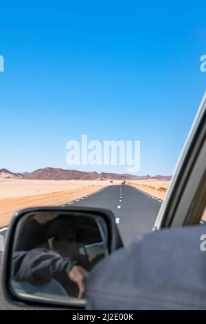 Lange Sahara Road Trip in leerer reg und sandiger Wüste, felsige Berge von der Rückseite aus gesehen ein fahrender Wagen, tuareg Fahrer im Spiegel nicht erkennbar. Stockfoto