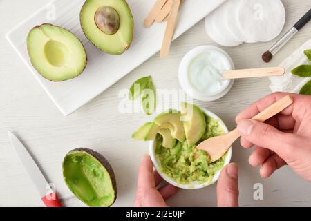 Hände, die natürliche Avocado-Hautpflegecreme vorbereiten, hergestellt in der Küche mit Fruchtstücken auf weißem Tisch und Küchenhintergrund. Horizontale Kompositio Stockfoto