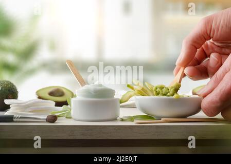Hände, die natürliche Avocado-Hautpflegecreme vorbereiten, hergestellt in der Küche mit Fruchtstücken auf weißem Tisch und Küchenhintergrund. Horizontale Kompositio Stockfoto
