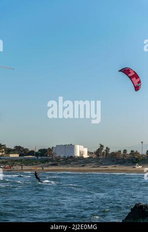 Strandsurfer hält Fallschirm in Meereswellen. Moretti, Hotel El Ryadh Building Tower. Tiefblaues Meer und Menschen am Sandstrand. Kiteboarding. Sidi Fredj. Stockfoto