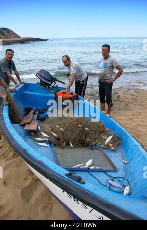 Fischer, die neben einem Boot aus dem Wasser stehen, voll mit Fischen, die im Fangnetz gefangen sind, paddeln. Ein Mann mit Tätowierungen im Arm, der einen Kanister hält. Stockfoto