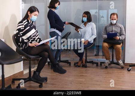 afroamerikanische Frau, die im Wartezimmer ein Jobangebot von der Schwangeren Managerin der Finanzabteilung erhält. Verschiedene Kandidaten im Lobbybereich lesen unternehmensinterne Vorschriften. Stockfoto