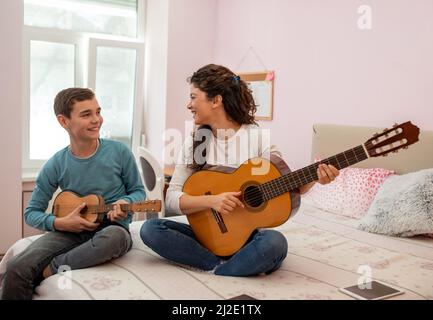 Mutter und Sohn sitzen auf einem Bett im Schlafzimmer und verbringen viel Zeit mit Spaß beim Spielen auf einer Gitarre. Stockfoto