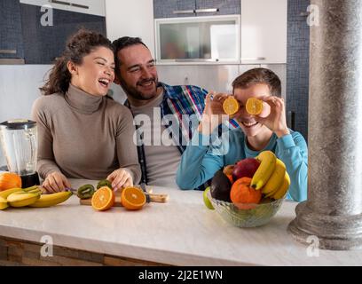 Mutter und Vater stehen in der Küche und lachen über ihren Sohn, der herumtollt, während er orangefarbene Scheiben auf die Augen hält Stockfoto