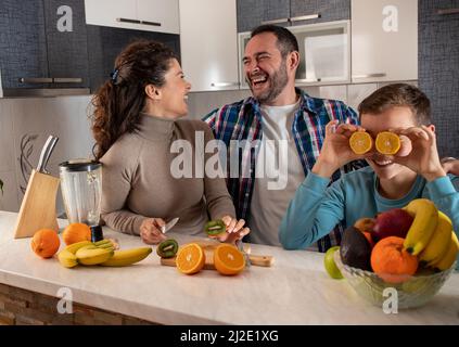 Mutter und Vater stehen in der Küche und lachen über ihren Sohn, der herumtollt, während er Orangen an den Augen hält Stockfoto
