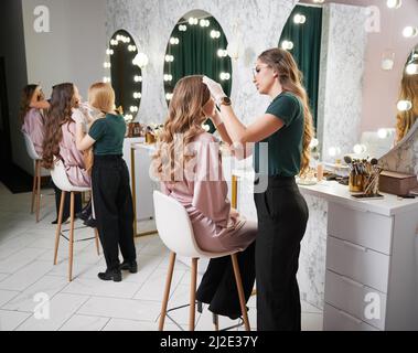 Beauty-Spezialisten tun professionelle Make-up für Kunden in modernen Salon. Junge Frau mit langen lockigen Haaren sitzt am Frisiertisch, während weibliche Stylistin Foundation mit Schwamm aufbringen. Stockfoto