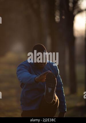 Porträt eines schwarzen Mannes in Sportkleidung, der sich an einem frühen Morgen in der Natur ausdehnt. Stockfoto