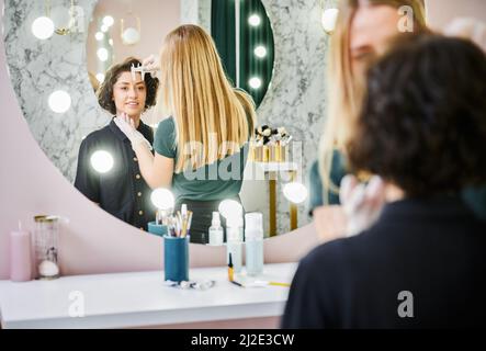 Junge Frau, die in den Spiegel schaut und lächelt, während der Ästhetiker die Augenbrauenentfernung mit dem Brauenlineal misst. Beauty-Spezialist mit Messwerkzeug vor Augenbrauenkorrektur Verfahren im Schönheitssalon. Stockfoto