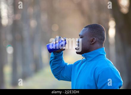 Der junge schwarze Mann in Sportkleidung trinkt nach dem Training aus einer Flasche Wasser und fühlt sich müde. Stockfoto