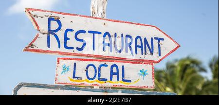 Ein Schild mit der Aufschrift „Lokale Restaurants“ zeigt auf der rechten Seite in einem tropischen Sommerziel auf einer rustikalen weißen Holztafel. Stockfoto