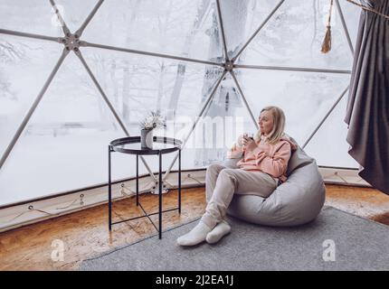 Eine Frau sitzt in einem Geo Dome Glamping Zelt in einem Taschenstuhl und blickt auf die schneebedeckte Winterlandschaft und trinkt Kaffee. Glamping Urlaub l Stockfoto