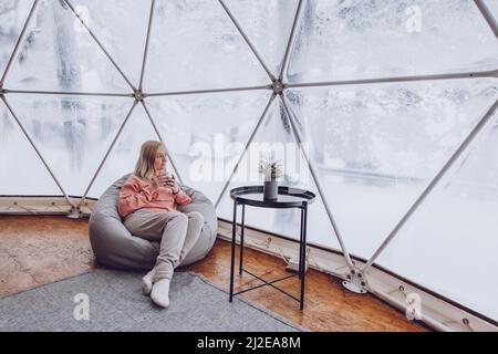 Eine Frau sitzt in einem Geo Dome Glamping Zelt in einem Taschenstuhl und blickt auf die schneebedeckte Winterlandschaft und trinkt Kaffee. Glamping Urlaub l Stockfoto