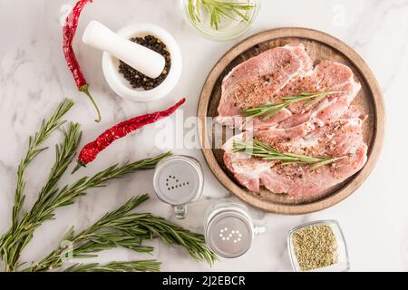 Zwei rohe Schweinesteaks auf einem runden Holztablett und ein Tisch aus weißem Marmor, der zum Grillen bereit ist Stockfoto