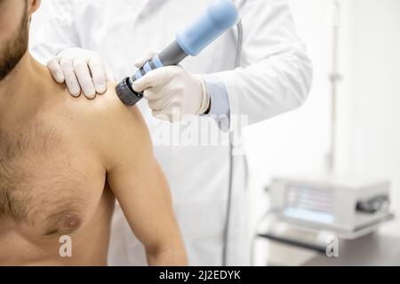 Der Arzt wendet eine Schockwellentherapie auf die Schulter des Mannes an Stockfoto
