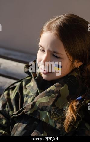 Trauriges Kind mit gemalter ukrainischer Flagge auf Wange und Tarnjacke, Stockbild Stockfoto