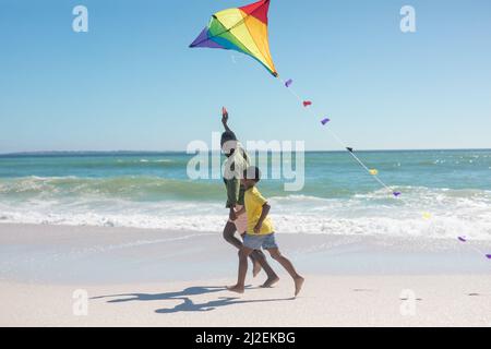 afroamerikanischer Vater und Sohn halten sich die Hände, während sie am sonnigen Tag mit Drachen am Strand laufen Stockfoto