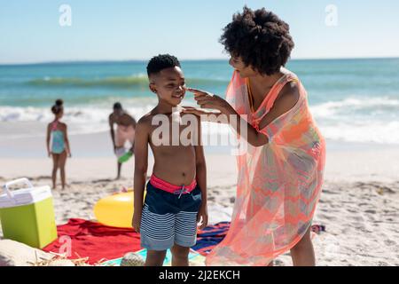 afroamerikanische Frau, die Lotion auf das Gesicht des Sohnes aufgibt, während Vater und Tochter am Strand spielen Stockfoto