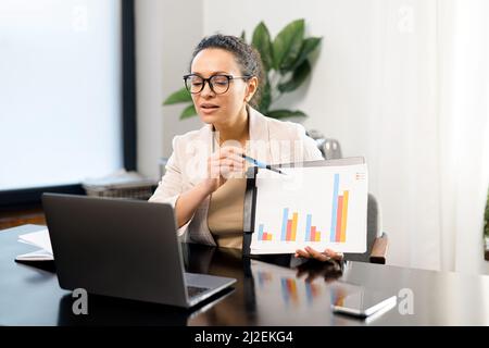 Virtuelle Konferenz. Weibliche Büroangestellte, die vor dem Laptop am Schreibtisch sitzt und an der Webcam ein Blatt mit Diagramm zeigt, und selbstbewusste Frau, die Ergebnisse berichtet. Business Coach hält Webinar ab Stockfoto