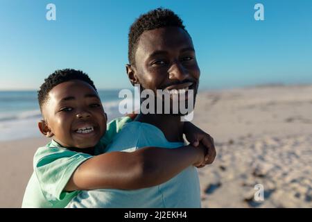 Porträt eines glücklichen afroamerikanischen Vaters Huckepack-Sohnes am Strand an einem sonnigen Tag Stockfoto