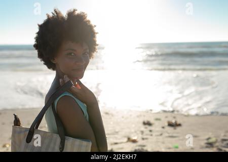 Seitenansicht Porträt einer lächelnden afroamerikanischen Frau mit Tasche am Strand an sonnigen Tagen Stockfoto